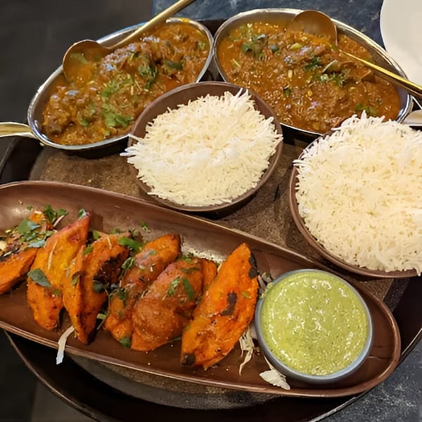 Two bowls of Indian curry, two bowls of rice, and a plate of roasted sweet potatoes with green sauce.