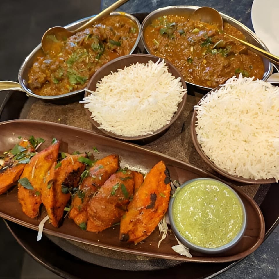Two bowls of Indian curry, two bowls of rice, and a plate of roasted sweet potatoes with green sauce.
