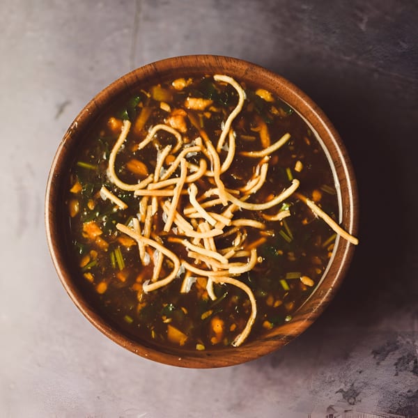 Overhead view of a wooden bowl filled with Indian hot and sour soup, topped with crispy noodles.